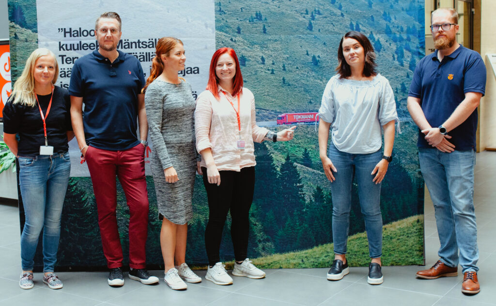 Six happy Tokmanni employees in front of the rollup wall